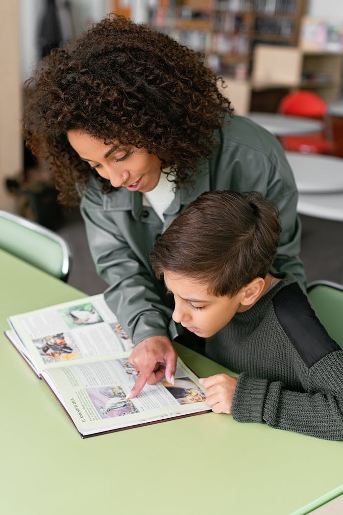 A teacher helps a student read a book at a green table in a classroom, fostering learning.